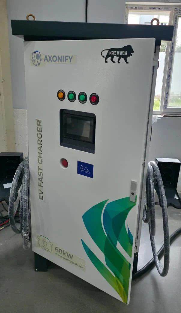 A white AXONIFY cabinet with a visible 60 kW marking and two charging cables. The photograph documents the equipment appearance; it does not establish commissioning status or Vasudha manufacturing ownership.